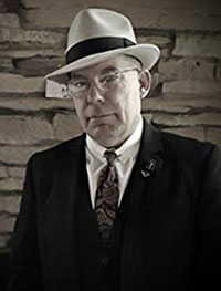 Headshot of an older white man wearing a white fedora and a black suit.