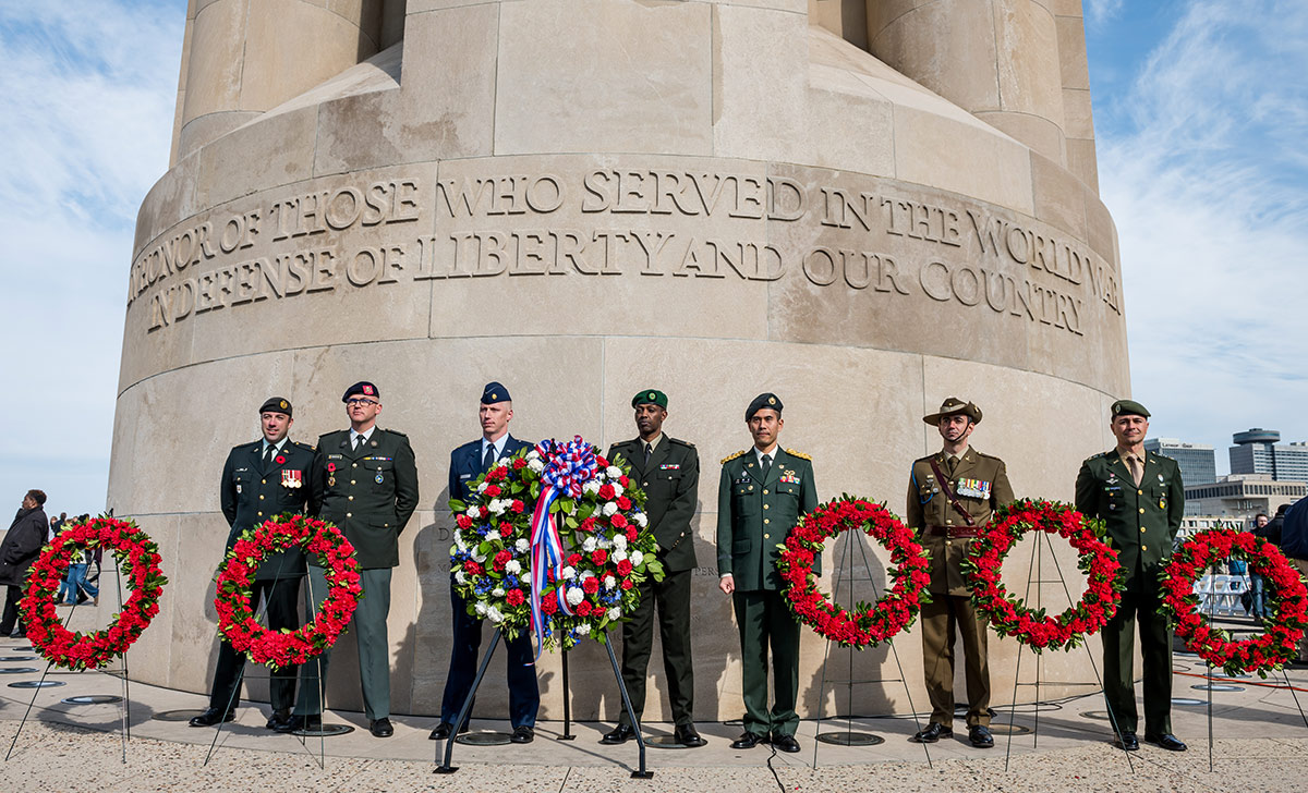 Modern photograph of men dressed in uniforms from different countries standing in a line behind a line of wreaths, all in front of the base of the Liberty Memorial.