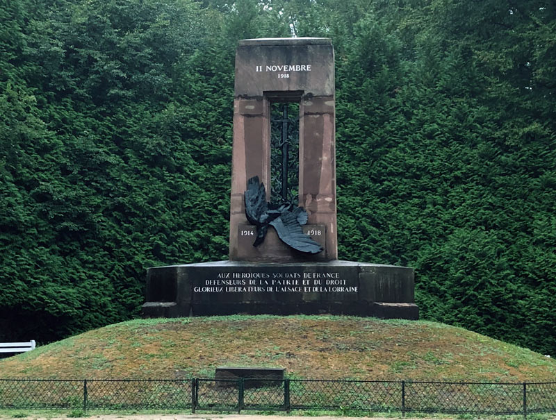 Photograph of a rectangular stone war memorial on a small hill. Text on top: 11 Novembre 1918. A statue of a fallen eagle with a sword hung over it is in the center of the rectangle. The memorial and hill are surrounded by thick green trees.