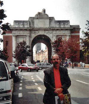 Photograph of a man with a mustache standing in front of a large European arch.