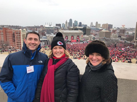 Photograph of a white man and two white women dressed in modern cold-weather jackets and hats posing on the Memorial courtyard terrace with a large crowd in the background.