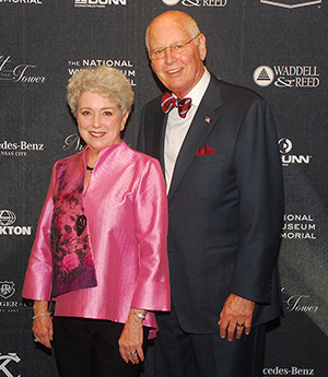 Modern photograph of an older white couple in front of a black photo backdrop. The woman is in a bright pink silk jacket. The man is in a black suit with a red and blue striped bowtie.