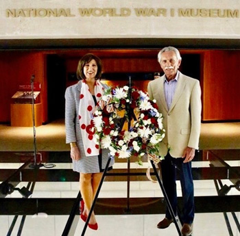 Modern photograph of a middle-aged white couple standing on the glass bridge of the Museum with a flower wreath between them.