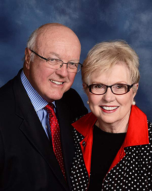 Portrait photograph of an older white couple against a blue backdrop. The woman has short blonde hair and wears a black and white polka-dot jacket with a red collar. The man wears a black suit with a red tie.