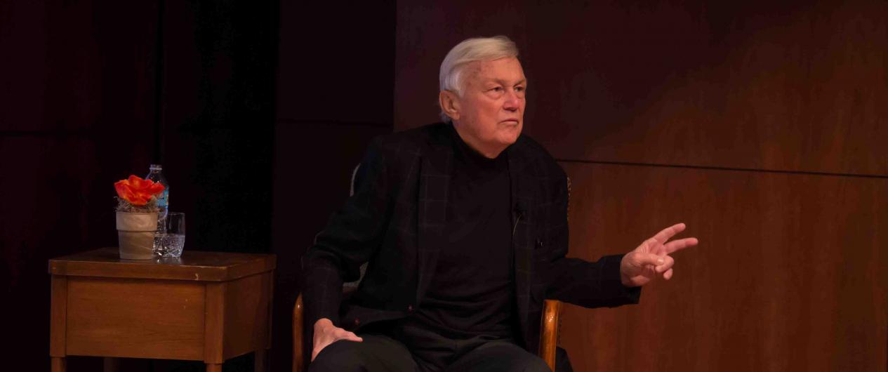 Photograph of an old white man with short silver hair dressed in a black suit and shirt. He is seated in a chair on the stage of the Auditorium.