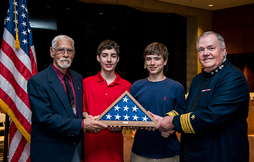 Two adult white men and two young white men hold a case containing a folded U.S. flag.