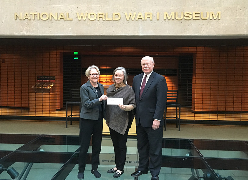 Photograph of an older woman, a younger woman, and an older man standing on the glass bridge of the Museum holding up a check.