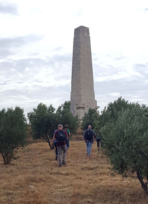 Several people in hiking gear walk towards a stone obelisk.