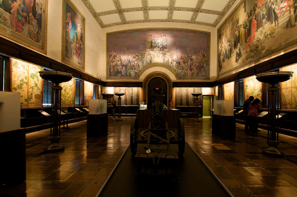 Modern photograph of the interior of a museum exhibit hall paneled in dark wood. The floor is dark stone. Lighted maps line the walls at head height. Above the maps the Pantheon de la Guerre mural dominates the walls.