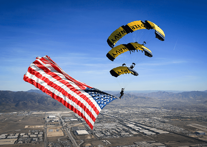 Multiple people gliding through the air on multiple parachutes. One has a giant U.S. flag attached to their feet so it streams out in the wind.