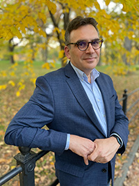 Portrait photograph of a middle-aged white man with dark hair wearing glasses and a blue blazer, leaning against a fence in a wooded area with golden leaves.