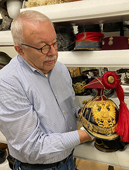 Modern photograph of an older white man with a white mustache and eyeglasses holding an elaborate WWI-era helmet