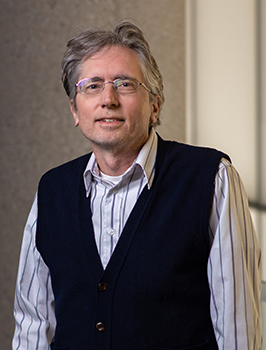 Modern photograph of an older white man with greying hair wearing glasses and a dark blue sweater vest.
