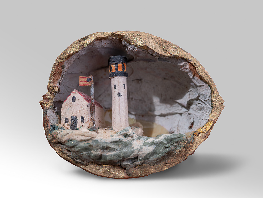 Modern photograph of a handcarved walnut shell diorama depicting a lighthouse, a cabin next to it, and a U.S. flag flying above the cabin.