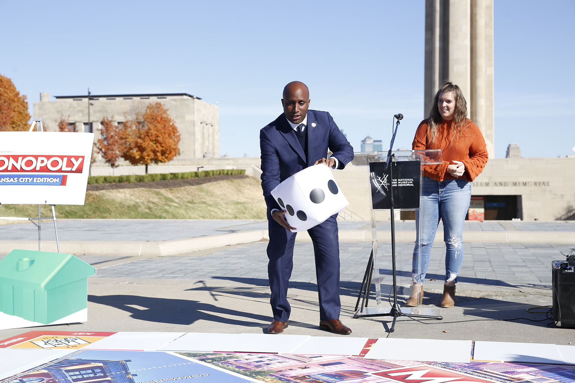 Modern photograph of Mayer Quinton Lucas, a Black man wearing a dark grey suit, about to roll an oversized die across an oversized game board in front of the Liberty Memorial Tower.