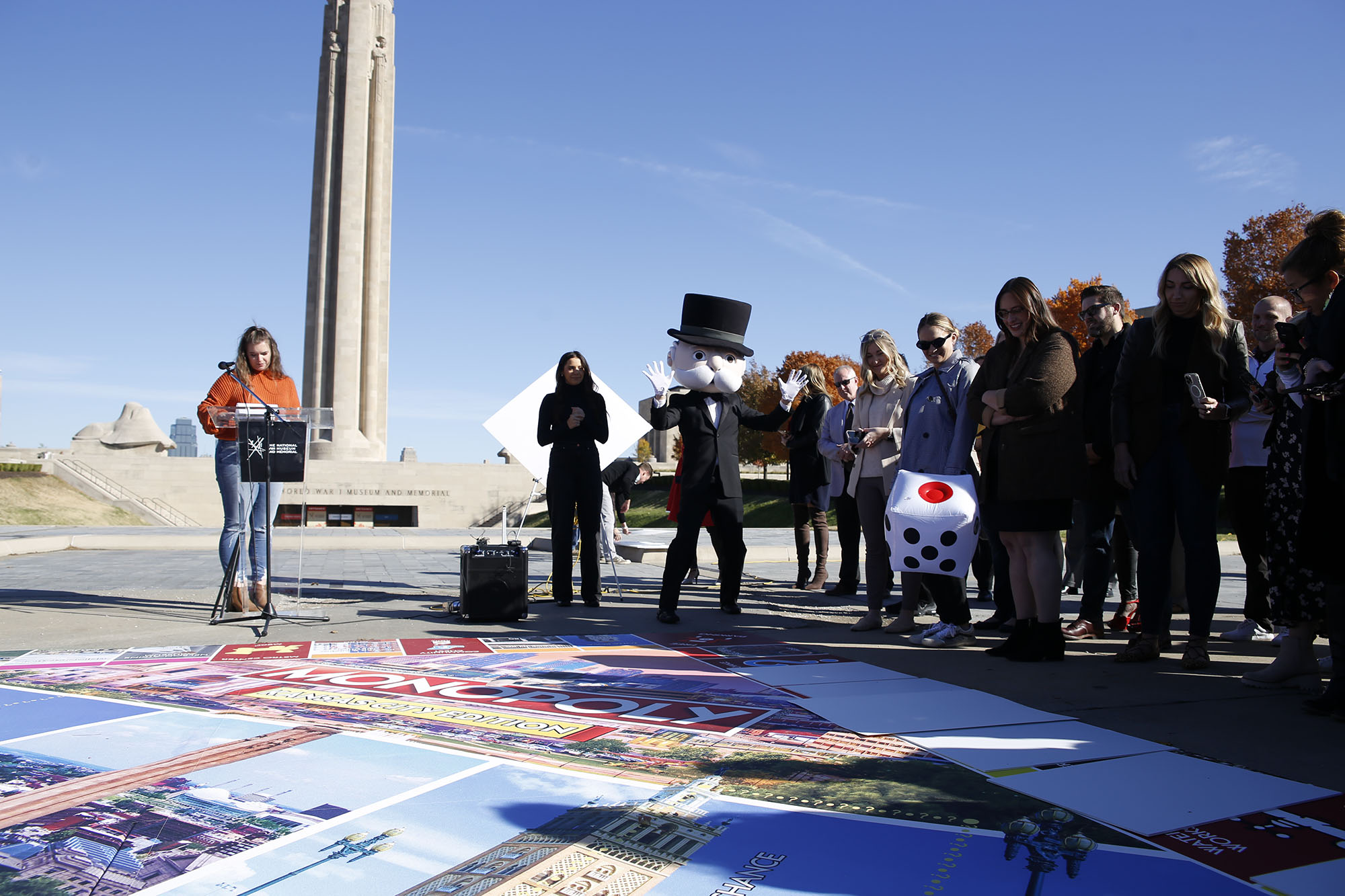 Modern photograph of an oversized version of a Monopoly board laid on the sidewalk in front of the Liberty Memorial Tower. Mr. Monopoly rolls an oversized die in front of a crowd.