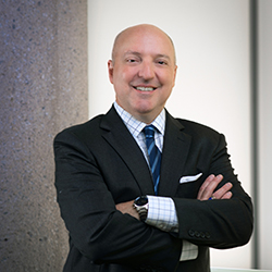 Headshot of a white man with a shaved head wearing a black suit and blue tie with his arms folded.