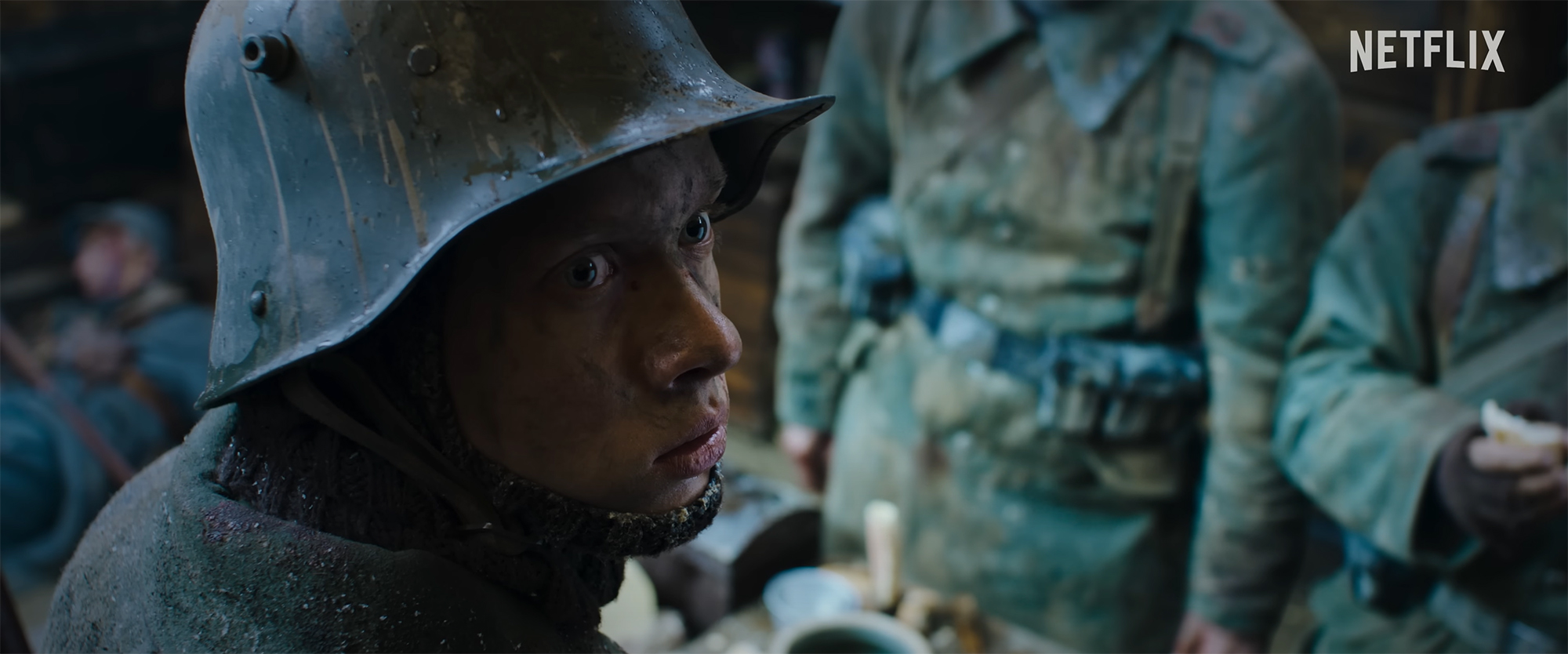 Film still depicting a young white man looking up and over his shoulder while wearing a round steel helmet