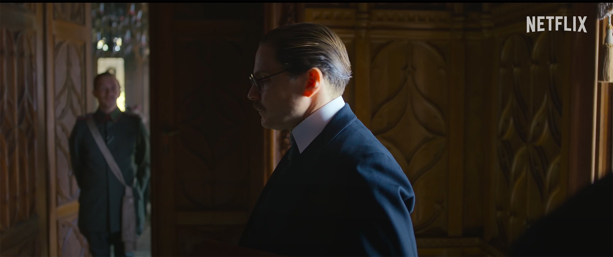 Film still showing a middle-aged man in a room with opulently carved wood walls, wearing a civilian suit. Another man wearing a military uniform stands in the door of the room.