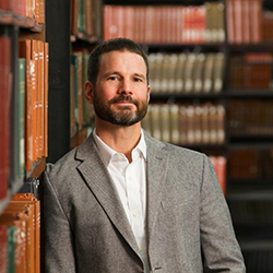 Tête d'un homme blanc avec une barbe et des cheveux noirs coupés court portant une veste de costume grise et debout dans une bibliothèque.