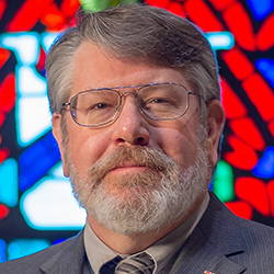 Headshot of a bearded white man with grey hair wearing glasses and a grey suit.