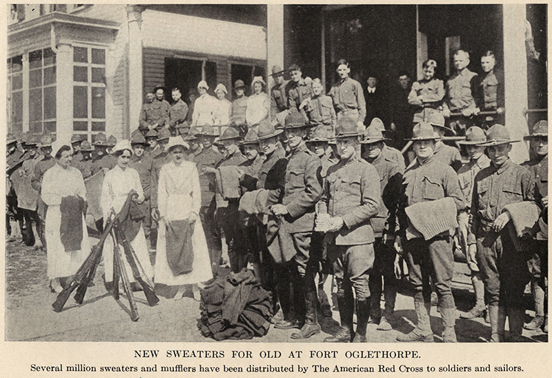Black and white photograph of a group of WWI soldiers standing with several Red Cross nurses, many of them holding knitted objects in their hands.