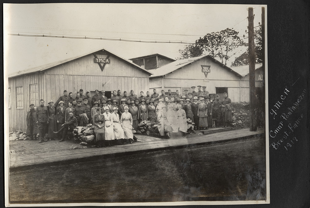 Black and white photograph of a large group of women and men in various uniforms gathered for a portrait in front of several wooden buildings adorned with YMCA signs.
