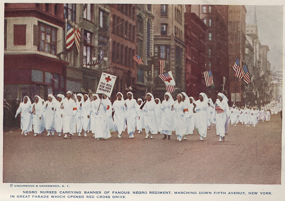 Color photograph of a group of Black women dressed in white nurse uniforms holding signs and walking down a street as part of a parade