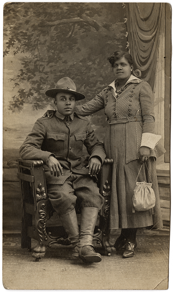 Sepia portrait photograph of a Black woman in a dress standing next to a seated Black man in WWI uniform