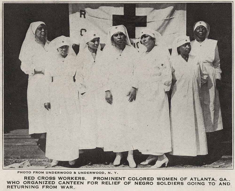 Black and white photograph of a group of Black women dressed in white nurse uniforms standing posed for a portrait with the Red Cross flag behind them.