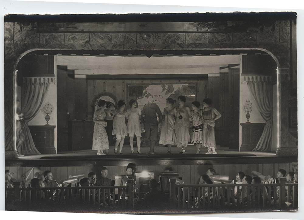 Black and white photograph of a WWI-era stage. A person dressed in a male WWI uniform is flanked by seven people dressed in a variety of frothy frocks.