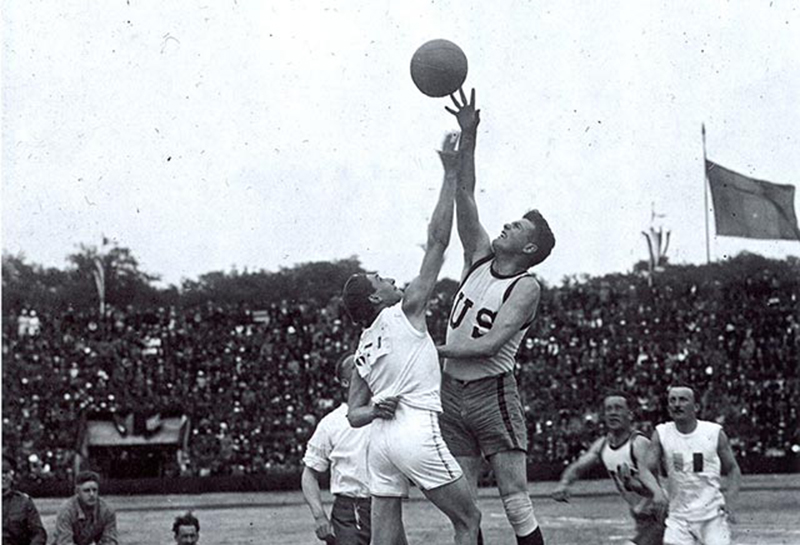 Black and white photograph of two white men wearing basketball uniforms jumping up in the air after a basketball