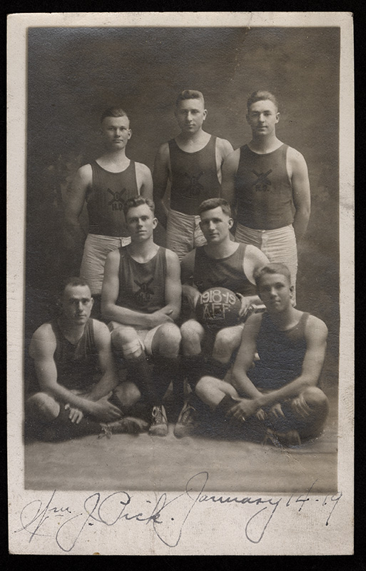 Photographie en noir et blanc de sept jeunes hommes blancs posant pour une photo de groupe (assis et debout) vêtus de shorts, débardeurs, chaussettes longues et baskets. L'un des hommes assis tient un ballon de basket sur ses genoux.