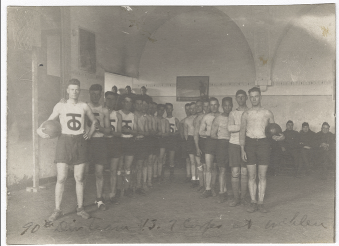Black and white photograph of eighteen young white men dressed in shorts, tank tops and sneakers posed in a V formation facing the viewer. Two are holding basketballs. Several more young men in military uniform sit on benches in the background of the gymnasium.