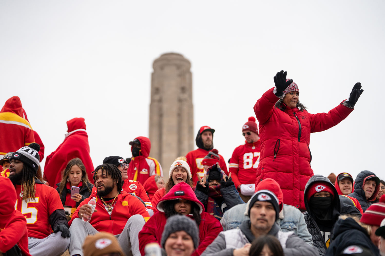 Modern photograph of a crowd of people dressed in red winter clothing. A person in a red puffy coat is standing up with her arms raised. The Liberty Memorial Tower is in the background.
