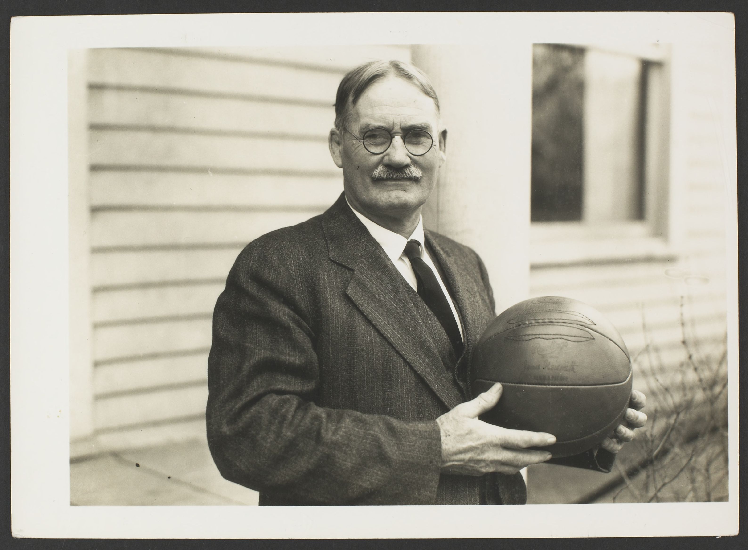 Sepia photograph of a man with a mustache and wire-framed glasses, wearing a suit and holding a basketball