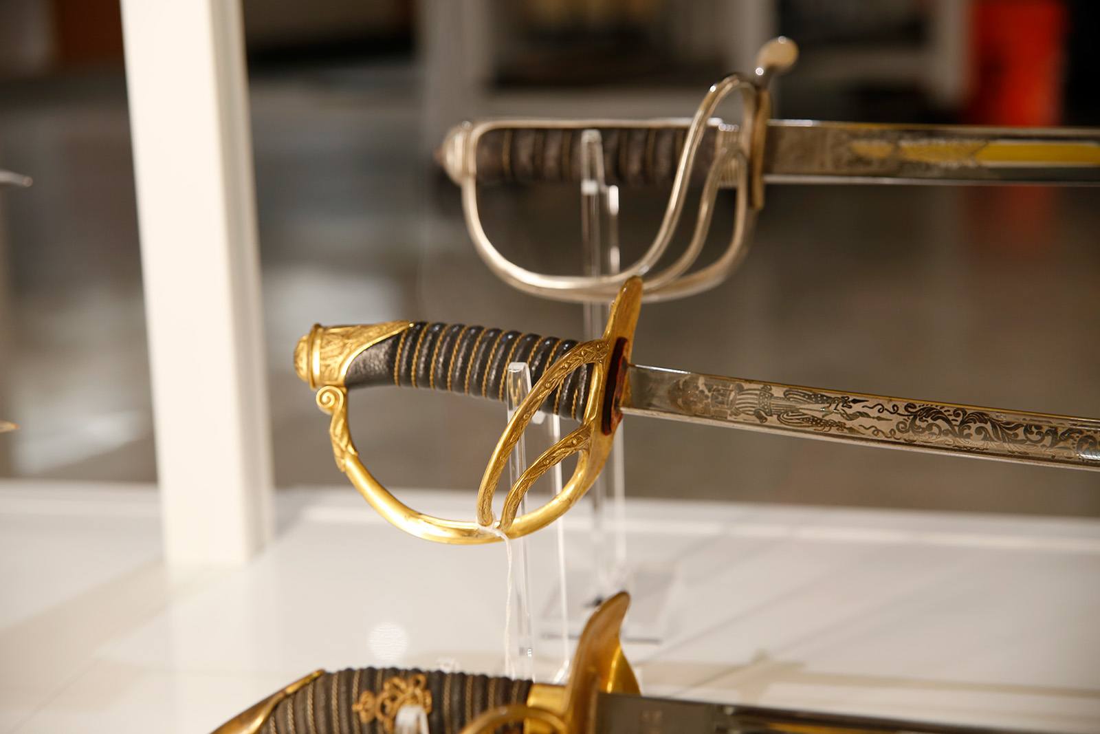 Modern photograph of a glass display case featuring three swords - focusing on the artfully engraved blades and hilts