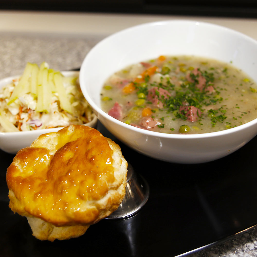 Modern photograph of stew in a bowl sitting on a tray with an American biscuit and a coleslaw salad in a small bowl