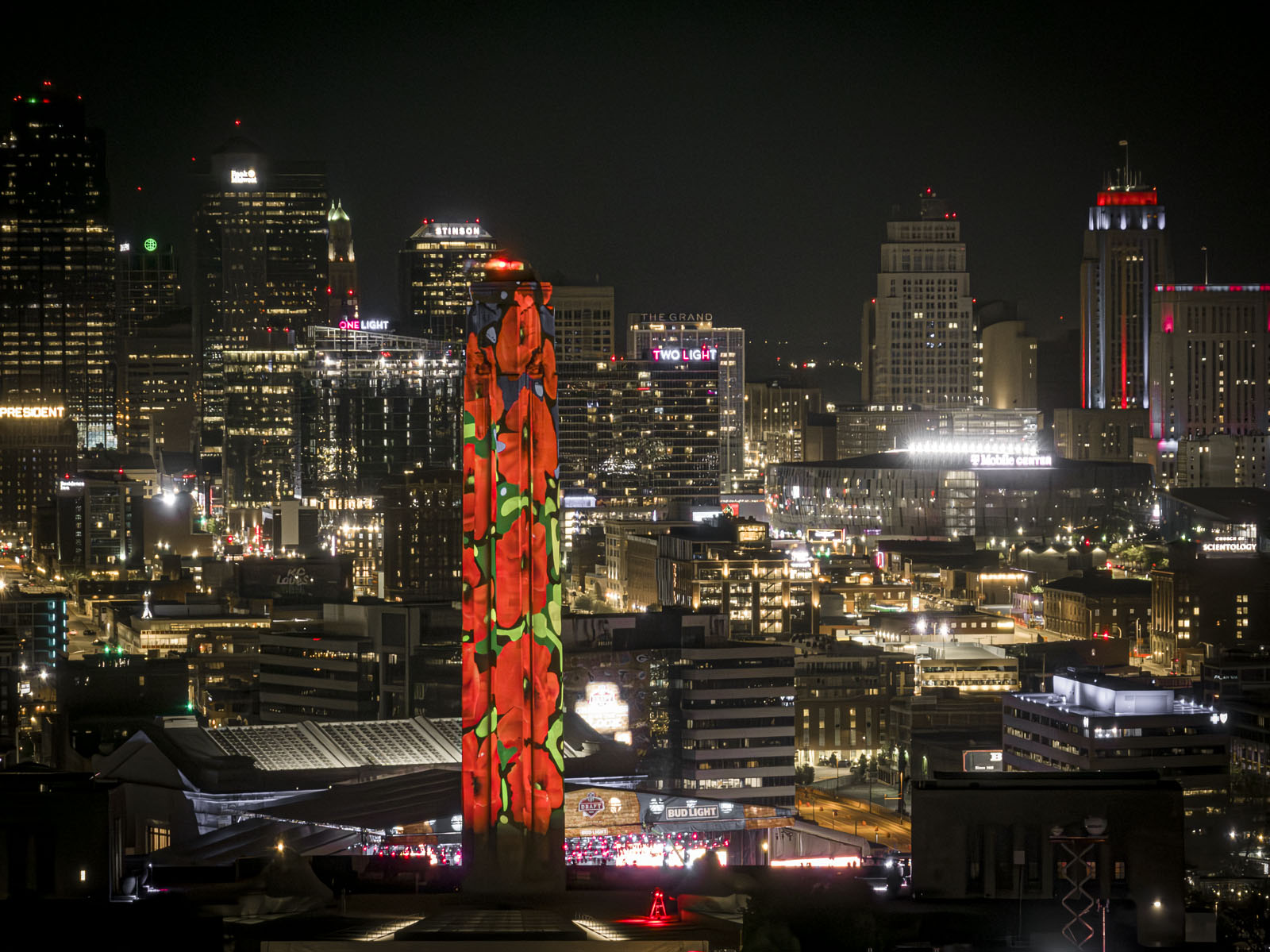 Modern photograph taken at night. Liberty Memorial Tower is in the foreground, lit up with poppy projections. The Kansas City downtown skyline is in the background, lit up brilliantly.