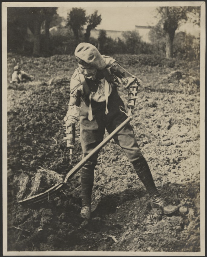 Black and white photograph of a French soldier using two prosthetic arms to move soil with a pitchfork. 