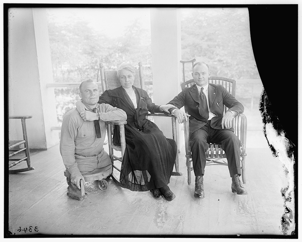 Black and white photo of three people posed for a group photo. Left to right: a white man in military uniform with both legs amputated nearly at the hip, resting on a small platform with wheels. A white woman in a dark-colored dress sitting in a rocking chair. A white man wearing a suit sitting in a rocking chair with a crutch leaning on a pillar behind him. The woman has her arm around the shoulders of the man on the left and her hand on the arm of the man on the right.
