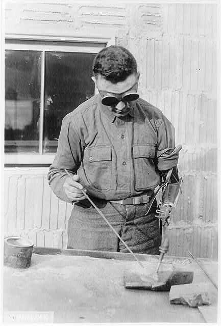 Black and white photo of a man wearing goggles and welding an object. His left arm is artificial.