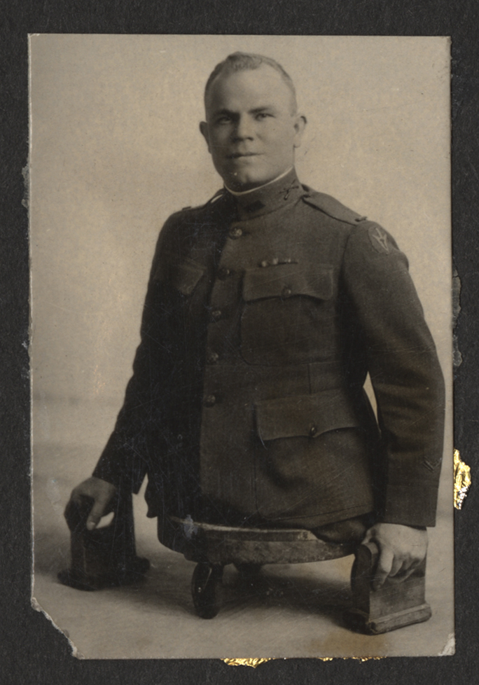 Black and white portrait photograph of an American soldier wearing military uniform. Soldier is missing both legs from the hips down. He sits on a flat, three-wheeled cart and holds blocks with handles to help him move. 