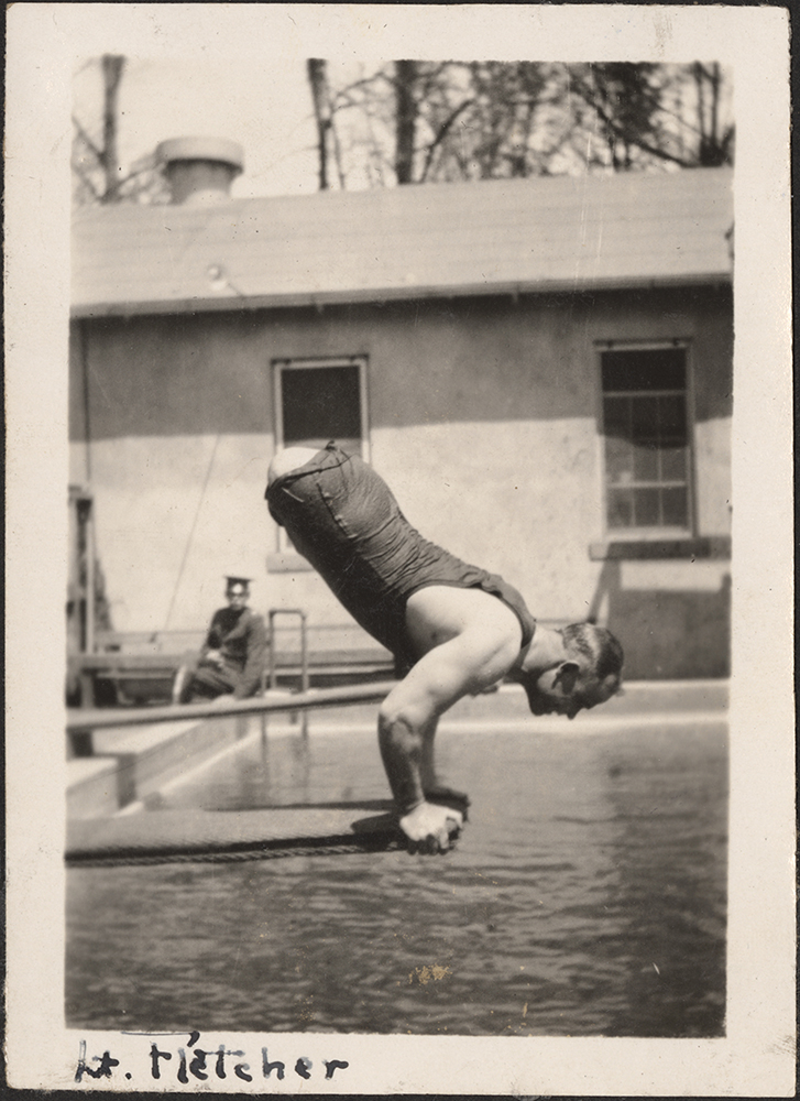 Black and white photograph of a white man (a double amputee) doing a handstand on a diving board over a swimming pool below. 