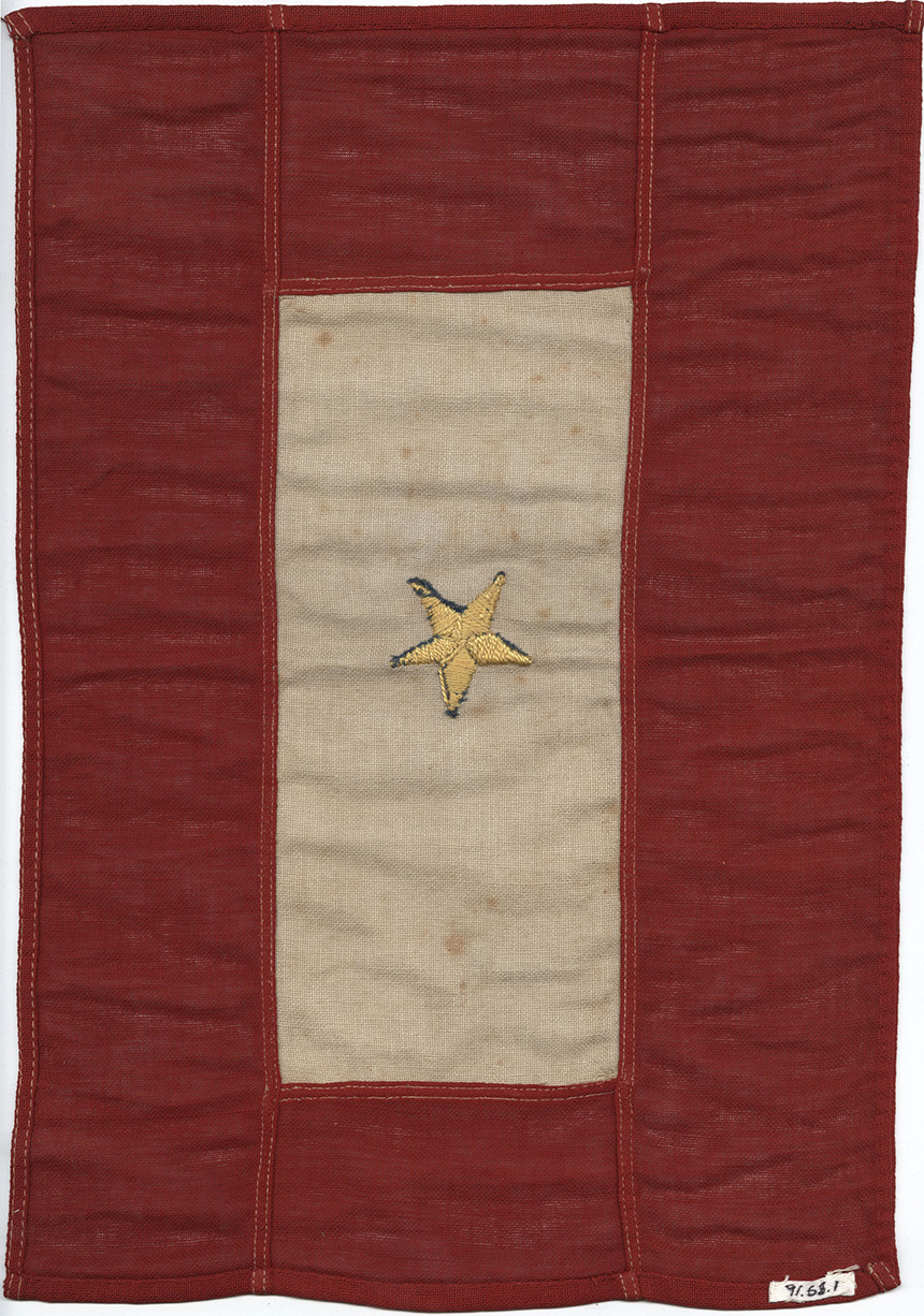 Modern photograph of a WWI-era flag. A red border surrounds a cream/white rectangle. A single gold star is embroidered in the center, with edges of blue showing that a blue star is underneath.