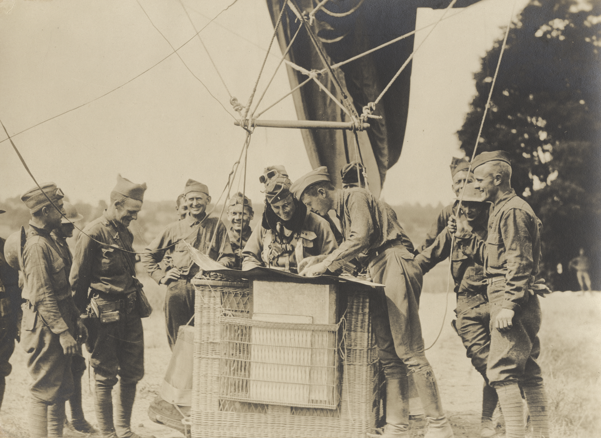 Black and white photograph of a WWI balloon basket tethered to the ground surrounded by smiling soldiers. The balloon pilot in the basket studies a large piece of paper.
