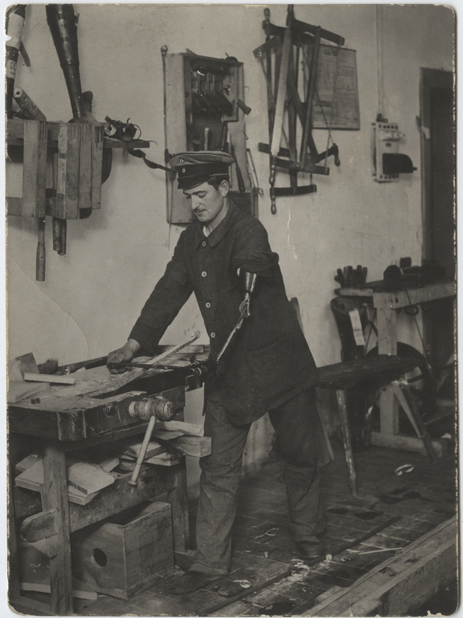 Black and white photograph of a man wearing a military cap and coat working in a woodshop. His left arm and hand are a jointed metal prosthesis.