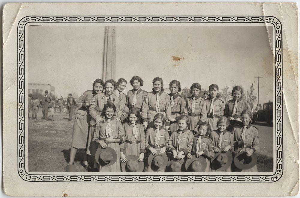 Full black and white photo of a group of girls and young women dressed in scouting uniforms posed with the Liberty Memorial Tower in the background. Some Boy Scouts are milling around behind them.