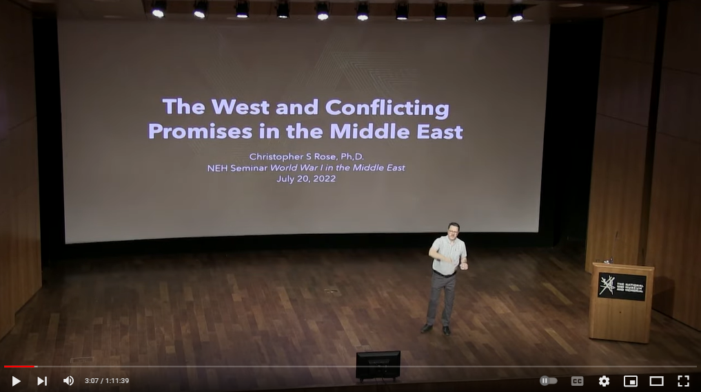 Screenshot of a lecture video. A white man speaks on a stage while a presentation is on the screen behind him.