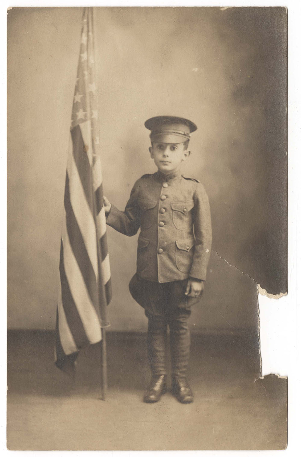 Photographie sépia d'un petit garçon blanc vêtu d'un uniforme militaire et d'une casquette, tenant un drapeau des États-Unis plus grand que lui.